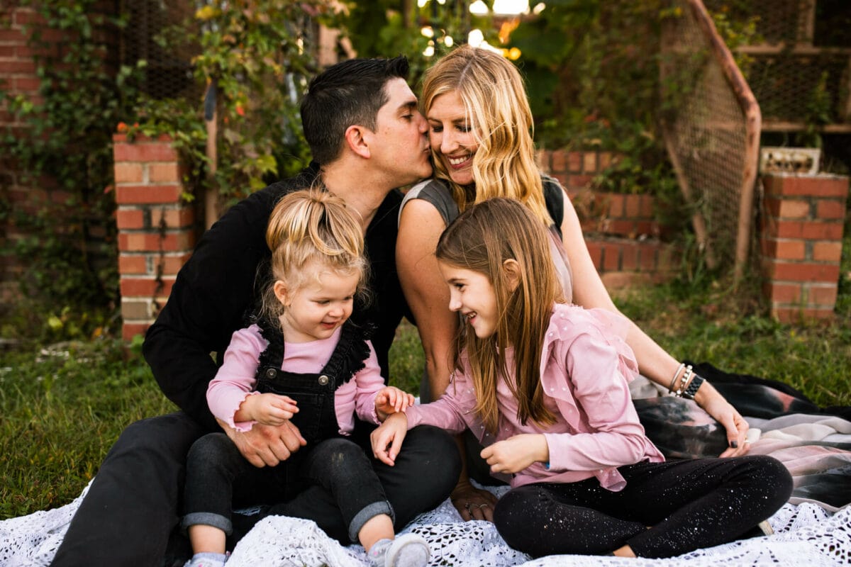 husband kisses wife while family is sitting on blanket