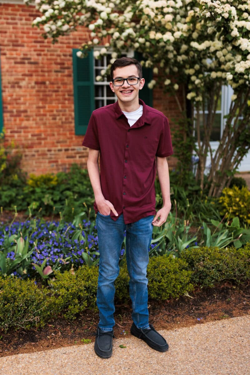 senior boy smiling in front of bush with flowers and hands in pockets